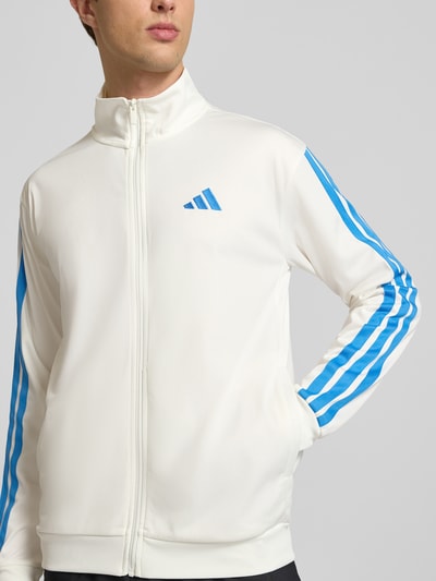 Männermodel trägt Regular Fit Trainingsjacke mit Stehkragen von adidas Sportswear, Optik: Unifarben, Passform: Regular-Fit, Verschluss: Reißverschluss, Logo-Print, Kontraststreifen, Kragenform: Stehkragen, Ärmelform: Langarm, Ärmelabschluss: Gerippt, Rumpfabschluss: Gerippt, Taschen: Seitliche Eingrifftaschen, Weiß mit blauen Kontraststreifen, glatte Textur, Ganzkörperansicht von vorne.