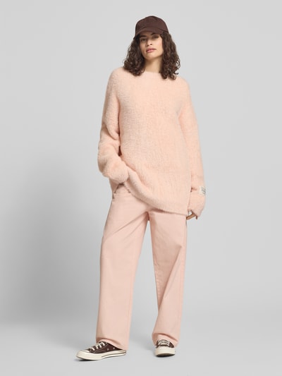 Frauenmodel trägt Oversized Strickpullover mit Label-Detail von Karo Kauer in Hellrosa, kombiniert mit weiter Hose und Cap, Ganzkörperansicht von vorne. Rückenlänge bei Größe S 76 cm, Ärmellänge 52 cm, Rundhalsausschnitt, überschnittene Schultern, weiche Textur.