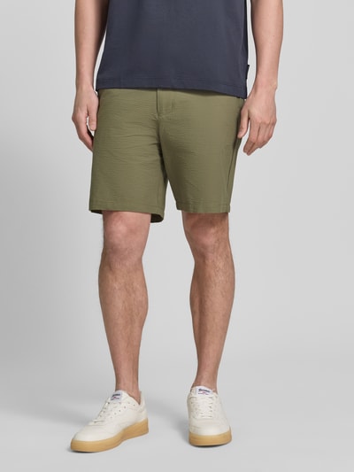 Männermodel trägt Regular Fit Bermudas aus Baumwoll-Mix Modell 'KARL' von SELECTED HOMME, wzór w paski, wykończenie materiału: seersucker, kolor oliwkowy, wpuszczane i tylne kieszenie, szlufki na pasek, zapięcie na guzik i zakryty zamek błyskawiczny, elastyczny ściągacz, krótka długość, zdjęcie: całkowity widok z przodu