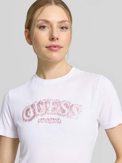Vrouwenmodel draagt T-Shirt uit viscose-elastaan mix met logo van Guess Activewear, regular fit, ronde hals, wit met roze logo, close-up bovenlichaam.