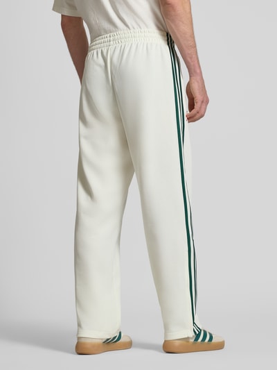 Männermodel trägt Sweatpants mit Reißverschlusstaschen und Logo-Stitching von adidas Originals, weiß mit grünen Streifen, Material-Mix, lockerer Schnitt, elastischer Bund, seitliche Reißverschlusstaschen, Ganzkörperansicht Rückansicht.