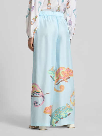 Frauenmodel trägt Wide Fit Seidenhose mit Motiv-Print von HERZENS in Hellblau mit bunten Chamäleon-Motiven, Elastikbund, Eingrifftaschen, Motiv-Print mit Tunnelzug, Reine Seide, Ganzkörperansicht Rückansicht.