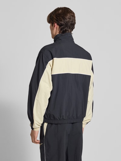 Männermodel trägt Trainingsjacke mit Logo und Reißverschluss von Nike im Regular-Fit mit Stehkragen, Rückenlänge 67 cm, Ärmellänge 64 cm, zweifarbig in Schwarz und Beige, glatte Textur, Ganzkörperansicht Rückansicht.