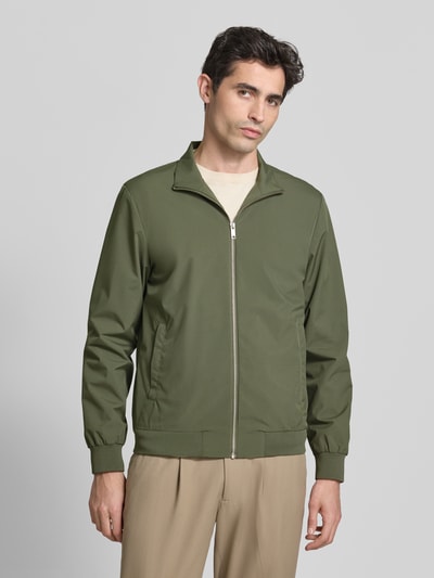 Mannelijk model draagt Jacke mit Stehkragen und Eingrifftaschen Model 'Joshu' van Casual Friday in olijfgroen met ritssluiting en steekzakken, regular fit, ruglengte maat M 68 cm, schouderbreedte 45 cm, borstwijdte 56 cm, mouwlengte 68 cm, voorkant geheel zichtbaar, Ganzkörperansicht von vorne.