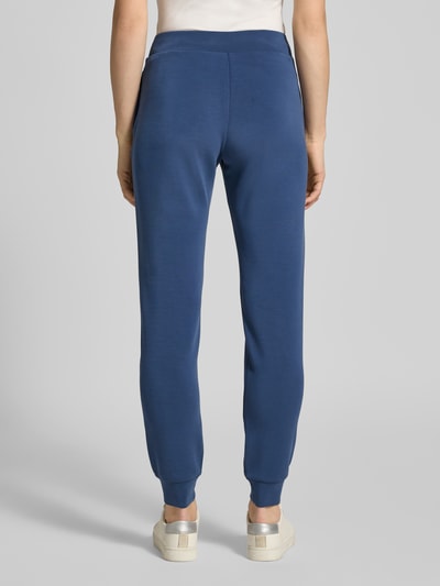 Kobiece modelka nosi Sweatpants mit Label-Detail od Guess Activewear, niebieski kolor, prosty krój, elastyczny pas, widoczne kieszenie boczne, materiał z dodatkiem modalu, długość nogawki wewnętrznej 79 cm w rozmiarze S, widok całej sylwetki z tyłu.