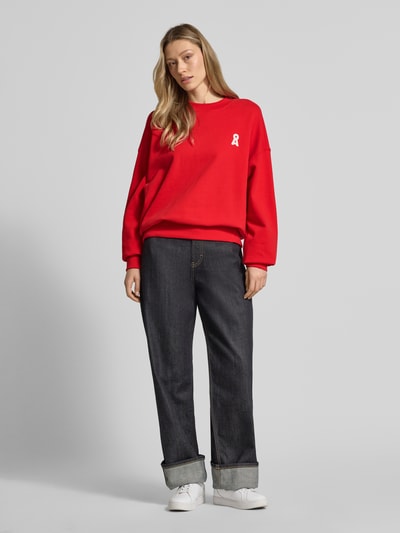 Frauenmodel trägt Oversized Sweatshirt aus reiner Baumwolle Modell 'ICONIC A ALIZAA' von Armedangels in Unifarbe Rot, Logo-Stitching, gerippte Abschlüsse, Crew Neck, Ganzkörperansicht von vorne.