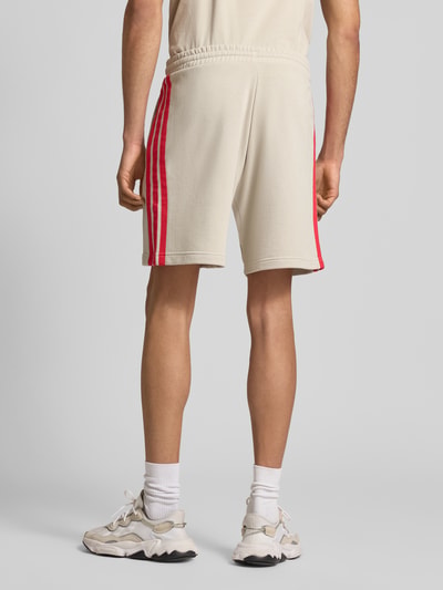 Männermodel trägt Regular Fit Sweatshorts aus Baumwoll-Mix von adidas Sportswear in Beige mit roten Kontraststreifen, Rückansicht Ganzkörperansicht.