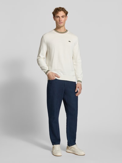 Männermodel trägt Regular Fit Pullover aus reiner Wolle von Lacoste in Creme mit dunkelblauer Hose, Ganzkörperansicht von vorne. Herren Pullover von Lacoste.