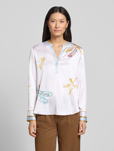 Kobiece model prezentuje Relaxed Fit Tunika-Bluse mit Allover-Motiv-Print od HERZENS, dekolt z lamówką, krój relaxed fit, nadruk z motywem na całej powierzchni, tył wydłużony, bluzka z długim rękawem, długość tyłu w rozmiarze 36: 68 cm, długość rękawów w rozmiarze 36: 63 cm, jasne kolory z żółtymi i niebieskimi detalami, gładka tekstura, widok całej sylwetki z przodu.