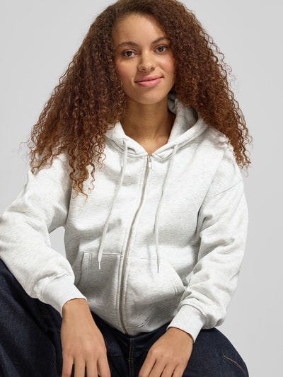 Modelka nosi Oversized Fit Sweatjacke mit geteilter Kängurutasche od Gina Tricot, mieszanka bawełny, kaptur z tunelem, elastyczne wykończenia, jasnoszara, gładka faktura, widok z przodu, zbliżenie na górną część ciała.