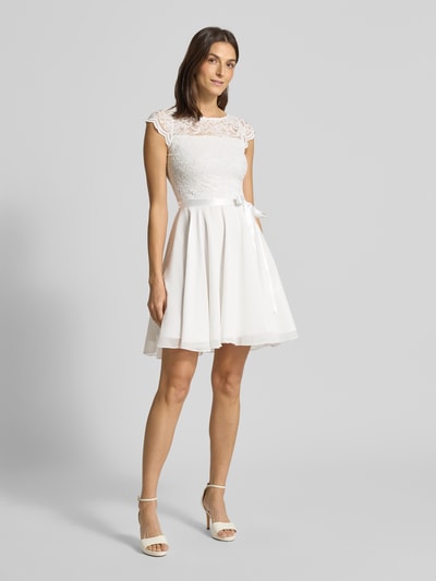 Frauenmodel trägt Cocktailkleid mit Spitzenbesatz und Bindegürtel von SWING, weißer Material-Mix mit Stretch-Anteil, A-Linie, Minikleid, Pailletten, Ganzkörperansicht von vorne. Rückenlänge 88 cm, Brustweite 39 cm, Größe 36.