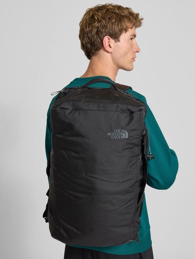 Männermodel trägt Duffel Bag mit Logo-Print Modell 'Voyager' von The North Face in Schwarz, strukturiertes Material, Nahaufnahme Oberkörper. Hauptfach mit Reißverschluss, verstellbare Schulterriemen, Innenfächer, Maße laut Kontext.