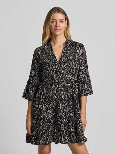 Kobiece modelka nosi sukienkę koszulową z wzorem na całej powierzchni, model 'EASY JOY' od Vero Moda, czarna z beżowym wzorem, rozkloszowana, długość mini, dekolt henley, marszczenia, rękaw o dł. 3/4, rozszerzane rękawy, prosty dół, materiał: czysta wiskoza; ujęcie: półzbliżenie przód