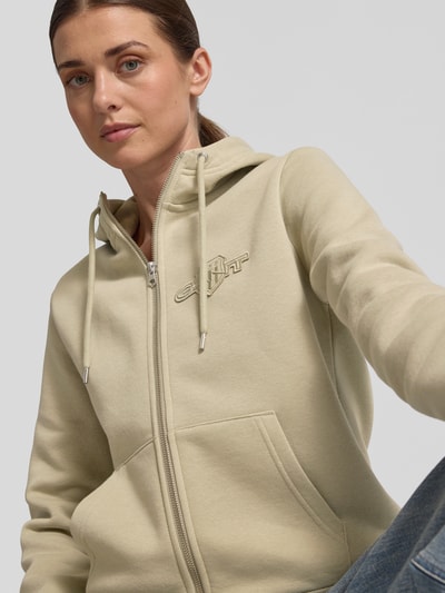 Frauenmodel trägt Sweatjacke mit Zweiwege-Reißverschluss und Label-Stitching von Gant, beige Baumwoll-Mix mit Känguru-Tasche, Nahaufnahme Oberkörper.