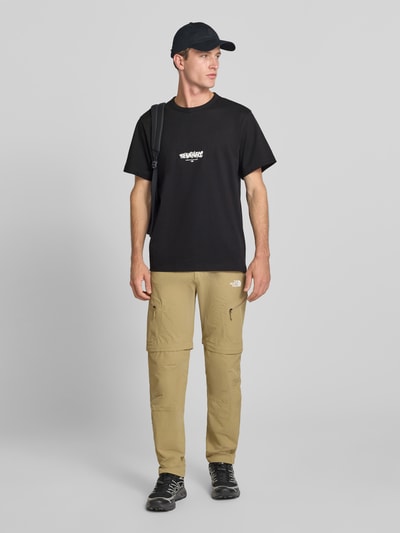 Männermodel trägt schwarzes Relaxed Fit T-Shirt aus reiner Baumwolle mit Logo-Print von The North Face, beige Hose, Ganzkörperansicht von vorne.