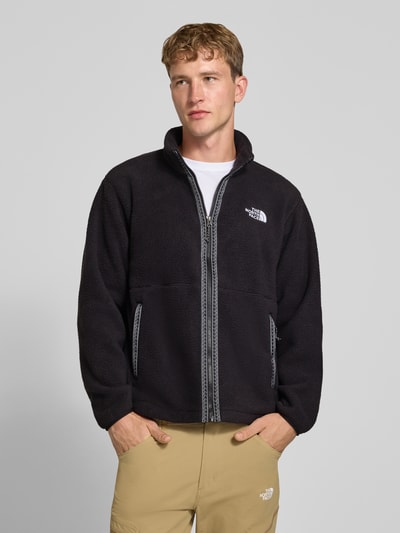 Männermodel trägt Regular Fit Fleecejacke mit Logo-Stitching von The North Face, schwarze Farbe, Reißverschluss, seitliche Eingrifftaschen, Stehkragen, Fleece-Textur, Ganzkörperansicht von vorne.