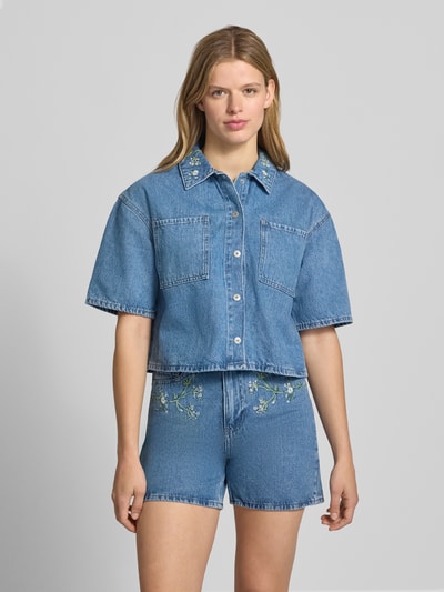Frauenmodel trägt Boxy Fit Jeansbluse aus reiner Baumwolle Modell 'SKY' von Pieces in Denim-Optik mit Stickerei, Kurzarm, Brusttasche, einreihige Knopfleiste, Rückenpasse, Cropped-Länge, Ganzkörperansicht von vorne.