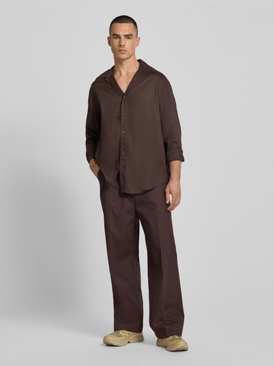 Männermodel trägt Wide Fit Hose aus reiner Baumwolle von Filippa K in Braun, mit Bügelfalten und Gesäßtaschen, Ganzkörperansicht von vorne.