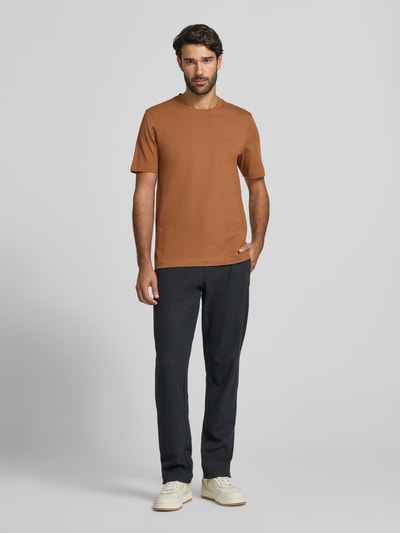 Mężczyzna nosi Regular Fit T-Shirt aus reiner Baumwolle von bugatti w kolorze brązowym, czarne spodnie, jasne buty, okrągły dekolt, wykończenia lamowane, długość tyłu w rozmiarze M: 69 cm, Ganzkörperansicht von vorne.