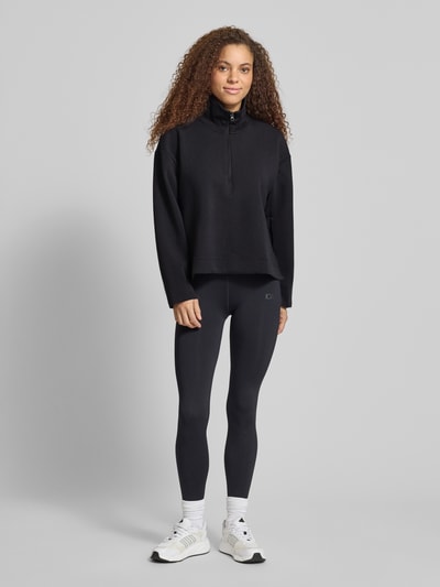 Frauenmodel trägt Relaxed Fit Sweatshirt mit Reißverschluss von adidas Sportswear in Schwarz, elastischer Material-Mix, Ganzkörperansicht von vorne.
