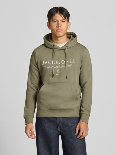 Männermodel trägt Relaxed Fit Hoodie aus Baumwoll-Mix Modell 'KYLE' von JACK & JONES in Olivgrün mit Känguru-Tasche, Ganzkörperansicht von vorne.