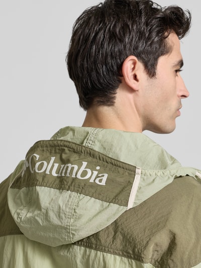 Männermodel trägt Windbreaker mit verdecktem Reißverschluss von Columbia in Grün mit Logo, unterschiedliche Texturen, Detailaufnahme Rückansicht, Regular-Fit, Kapuze mit Tunnelzug, Rückenlänge 70 cm, Schulterbreite 64 cm, Brustweite 57 cm, Ärmellänge 56 cm.