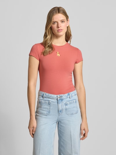 Kobiece model prezentuje damski t-shirt z mieszanki bawełny i elastanu, krój slim fit, wyhaftowany motyw, okrągły dekolt, długość tyłu 54 cm w rozmiarze S, T-Shirt mit Motiv-Stitching und Rundhalsausschnitt od REVIEW, jasnoróżowy kolor, gładka faktura, jasnoniebieskie jeansy, pełny widok z przodu.