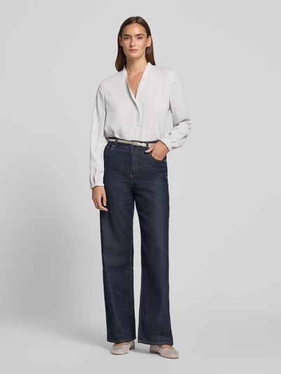 Kobiece modelka nosi Regular Fit Bluse mit V-Ausschnitt von Marc Cain, krój regular fit, dekolt w serek, detal z logo, długość tyłu w rozmiarze 36: 65 cm, długość rękawów w rozmiarze 36: 59 cm, jasna bluzka, ciemne spodnie, Ganzkörperansicht von vorne.