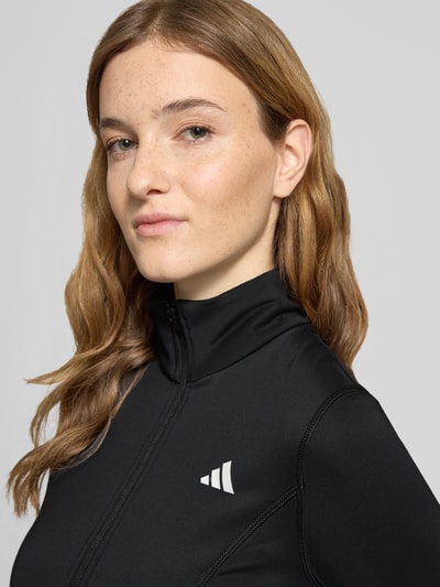 Frauenmodel trägt Slim Fit Jacke mit Stehkragen von adidas Sportswear, Schwarz, Logo-Print, elastischer Material-Mix, Nahaufnahme Oberkörper.