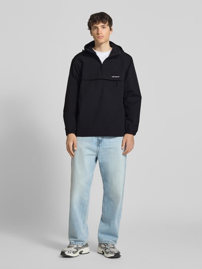Männermodel trägt Windbreaker mit Label-Stitching von Carhartt Work In Progress, schwarz, Kapuze, Reißverschluss, Regular-Fit, Ganzkörperansicht von vorne, Rückenlänge bei Größe M: 73 cm, Schulterbreite: 48 cm, Brustweite: 55 cm, Ärmellänge: 66 cm.