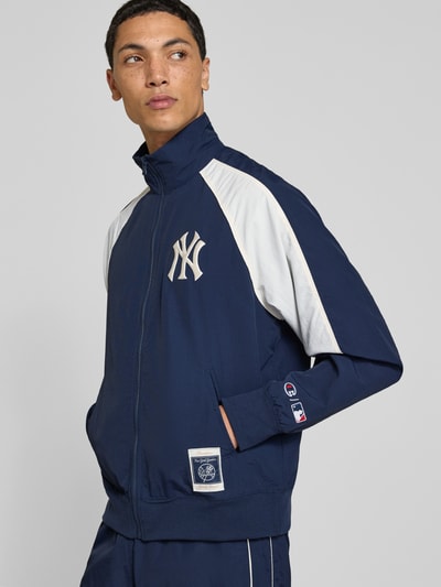 Männermodel trägt Relaxed Fit Tracksuit mit Logo-Patch von Champion in Dunkelblau mit weißen Einsätzen, Stehkragen, Reißverschluss, elastische Abschlüsse, Ganzkörperansicht von vorne, Herren Tracksuit, Logo-Patch, Bundweite 39 cm, Rückenlänge 68 cm, Schulterbreite 42 cm, Ärmellänge 49 cm, Innenbeinlänge 72 cm.