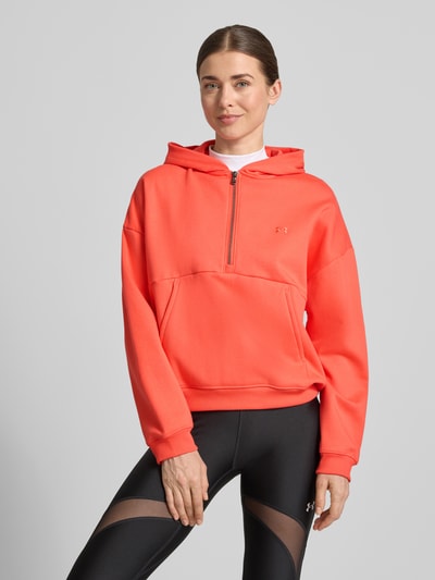 Modelka kobieca nosi Hoodie mit Reißverschluss und Label-Stitching od Under Armour, bawełniana mieszanka, krój regular fit, kieszeń kangurka, kolor koralowy, widok całej sylwetki z przodu.