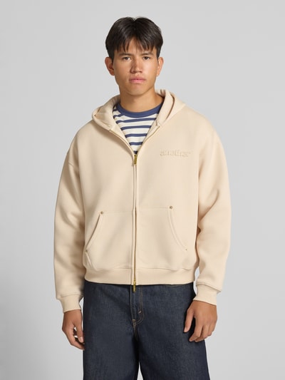 Männermodel trägt Sweatjacke mit Kapuze von ANOTHER COTTON LAB in Beige, lockerer Schnitt, Zwei-Wege-Reißverschluss, Känguru-Tasche, Ganzkörperansicht von vorne.