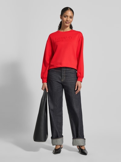 Frauenmodel trägt Sweatshirt mit Viskose-Anteil von monari in Rot, lockere Passform, Rundhalsausschnitt mit Nieten, Jeans, Ganzkörperansicht von vorne.