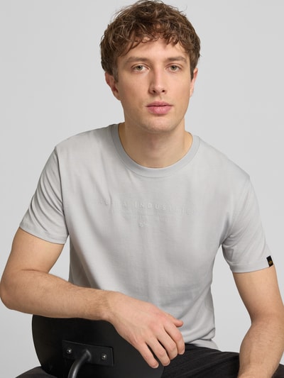 Męski model nosi T-Shirt mit Label-Print und Rundhalsausschnitt od Alpha Industries, bawełna, krój regular fit, długość tyłu w rozmiarze M 67 cm, jasnoszara, gładka faktura, widok na górną część tułowia z przodu.