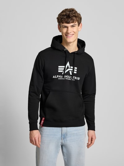 Männermodel trägt Hoodie mit Label-Print von Alpha Industries in Schwarz, Baumwollmischung, Känguru-Tasche, lockere Passform, Nahaufnahme Oberkörper.