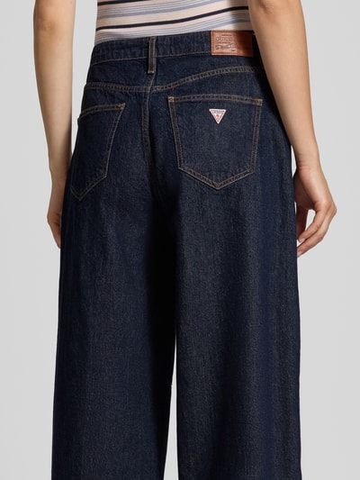 Frauenmodel trägt Barrel Fit Jeans in verkürzter Passform von GUESS, dunkles Denim, 5-Pocket-Design, Logo-Detail, Ganzkörperansicht Rückansicht.