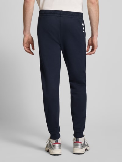 Männermodel trägt Sweatpants mit Logo und elastischem Bund von EA7 Emporio Armani in Dunkelblau, Regular Fit, Ganzkörperansicht Rückansicht.