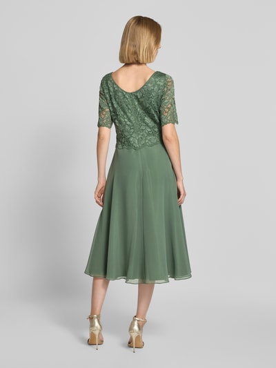 Frauenmodel trägt Tailliertes Cocktailkleid mit Spitzenbesatz von Vera Mont in grün mit floraler Struktur, Ganzkörperansicht Rückansicht.
