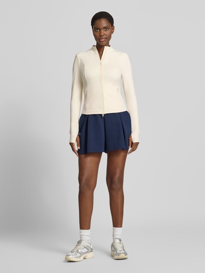 Frauenmodel trägt Jacke mit Zweiwege-Reißverschluss Modell 'Sense' von aim'n in Creme mit Stretch, kombiniert mit dunkelblauem Rock, Ganzkörperansicht von vorne. Slim Fit, Stehkragen, Reißverschlusstaschen, Maße laut Kontext.