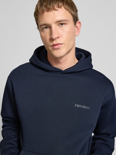 Männermodel trägt Regular Fit Hoodie mit Logo-Print von Review aus Baumwoll-Mix in Dunkelblau, Nahaufnahme Oberkörper.