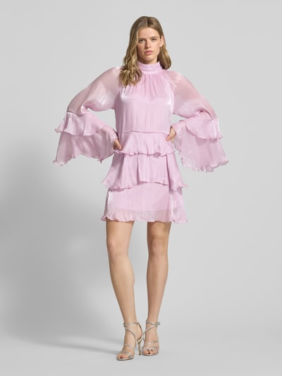 Frauenmodel trägt Minikleid im Stufen-Look Modell 'Flowy frill' von Gina Tricot in zartem Rosa mit Volants, transparenten Ärmeln und Stehkragen, A-Linie, rückseitige Schnürung, Ganzkörperansicht von vorne.