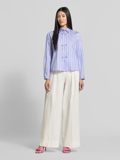 Frauenmodel trägt Regular Fit Hemdbluse aus reiner Baumwolle von Milano Italy in Blau mit rosa Streifen, Hemdblusenkragen, Zierschleifen, langärmliges Design, kombiniert mit weiter, heller Hose, Ganzkörperansicht von vorne.