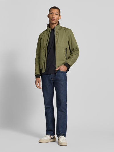 Männermodel trägt Regular Fit Jacke mit Stehkragen von MCNEAL in Oliv mit Reißverschluss, Eingrifftaschen, elastische Abschlüsse, Ganzkörperansicht von vorne. Rückenlänge 64 cm, Schulterbreite 43 cm, Brustweite 55 cm, Ärmellänge 67 cm bei Größe M.
