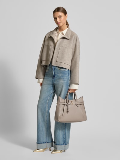 Frauenmodel trägt Handtasche in Leder-Optik mit Logo-Applikation von Guess in Taupe, strukturierte Oberfläche, silberne Details, Hauptfach mit Reißverschluss, Tragehenkel, abnehmbarer Schulterriemen, Innenfach mit Reißverschluss, Maße 40x25x13 cm, Schulterriemen 115 cm, Ganzkörperansicht von vorne.