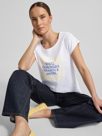 Frauenmodel trägt T-Shirt mit Statement-Print von Christian Berg Woman in Weiß mit gelb-blauem Print, Reine Baumwolle, Regular-Fit, Rundhalsausschnitt, Rollsaum, Rückenlänge 60 cm bei Größe S, Ganzkörperansicht von vorne.