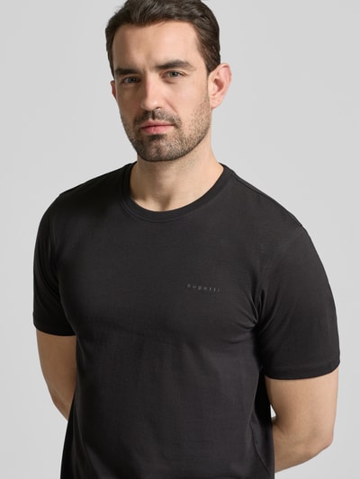 Männermodel trägt T-Shirt mit Label-Detail von bugatti, schwarz, reine Baumwolle, gerader Schnitt, Rundhalsausschnitt, Nahaufnahme Oberkörper.