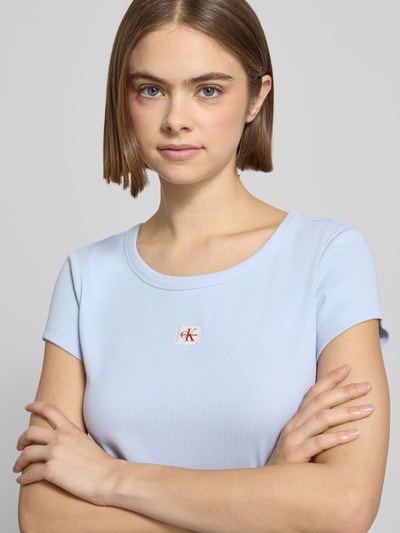 Kobiece model nosi Cropped T-Shirt mit Label-Detail od Calvin Klein Jeans, mieszanka bawełny i elastanu, okrągły dekolt, krótki krój, detail z logo, jasnoniebieski, damski t-shirt, długość tyłu 43 cm w rozmiarze S, zbliżenie na górną część ciała.