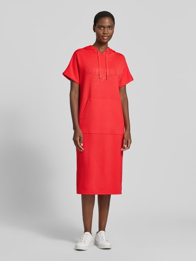 Kobiece model nosi Sweatkleid mit Kapuze od Soyaconcept, mieszanka materiałów z domieszką modalu, prosty krój, sukienka midi, kaptur, nadruk z logo, czerwony kolor, gładka faktura, widok całej sylwetki od przodu.