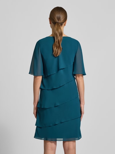Frauenmodel trägt Relaxed Fit Kleid mit Ziersteinbesatz von Christian Berg Cocktail in Blau, semitransparent, blickdichter Unterrock, Ganzkörperansicht Rückansicht.
